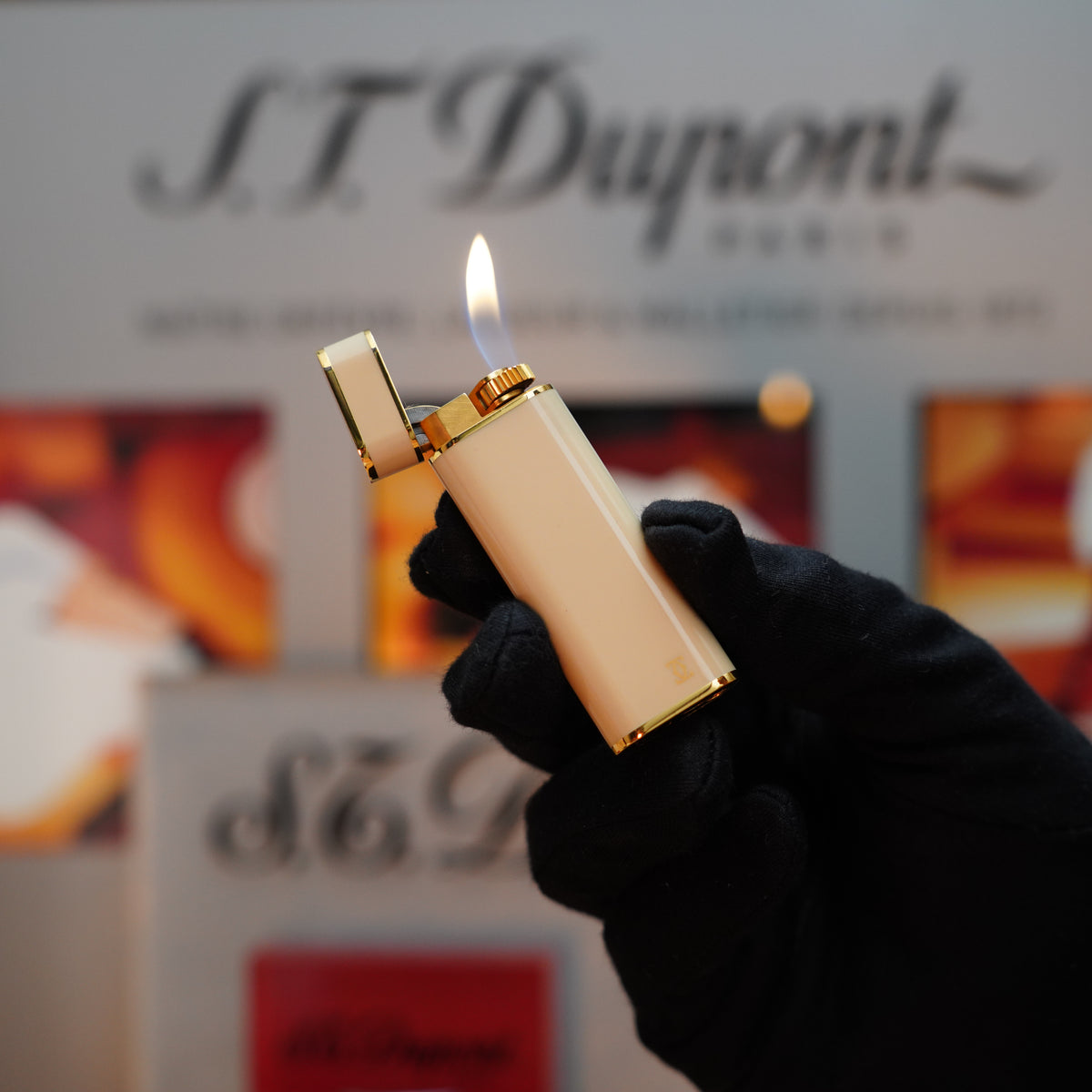 A gloved hand holds an elegant cream-colored Cartier lighter, specifically the Vintage 1980 Cartier Oval Le Must with a 24k gold finish and amber lacquer, its flame dancing against a blurred branded background.