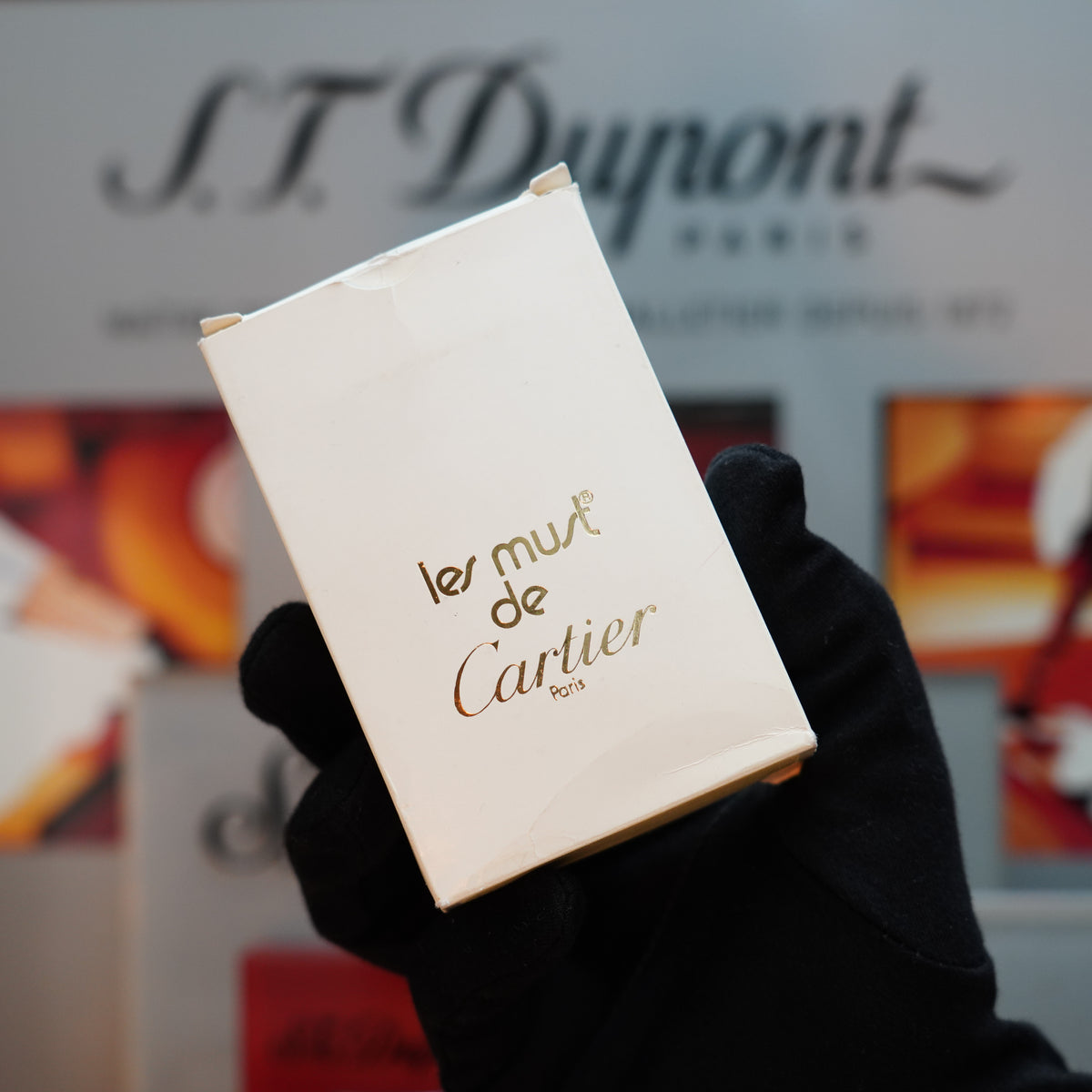 A gloved hand delicately holds a white box labeled "Les Must de Cartier," hinting at the elegance of the Vintage 1980 Cartier Oval 24k Gold Finish Amber Lacquer Le Must Lighter. In the blurred background, text reads "S.T. Dupont Paris," softly suggesting luxury similar to that of this exquisite Cartier lighter.