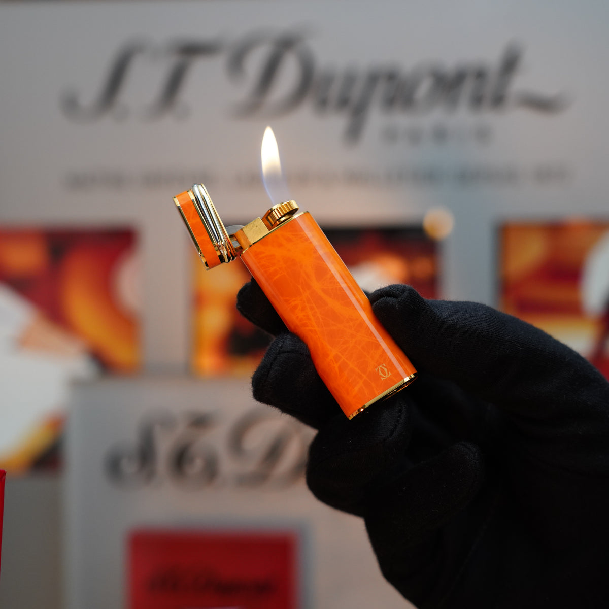 A gloved hand holds a Vintage 1980 Cartier Oval 24k Gold Finish Orange Marble Lacquer Lighter, its flame flickering mysteriously as blurred branding and logos create an enigmatic backdrop.