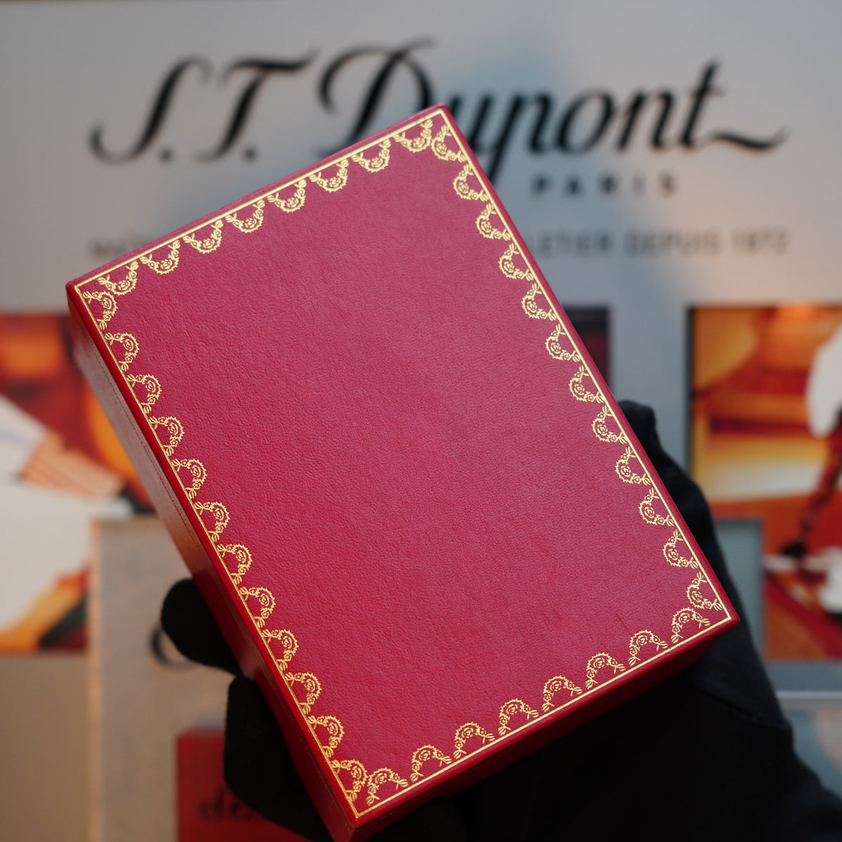 A person wearing a black glove is holding a red box with gold decorative edging, reminiscent of the Vintage 1980 Cartier design. In the background, the blurred Cartier sign adds an air of elegance to this vintage-inspired setting.