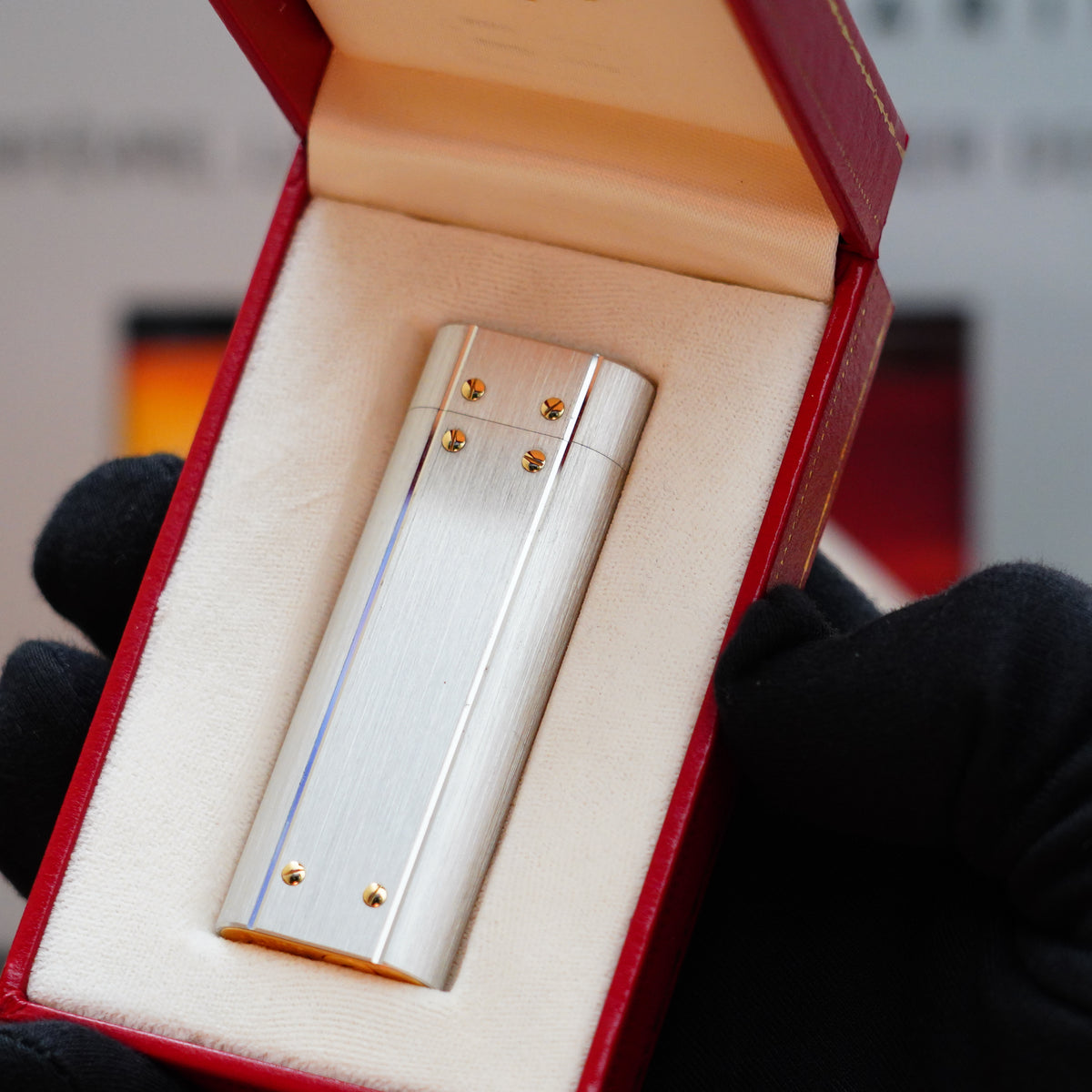 A hand in a black glove holds a Vintage 1982 Cartier Santos Palladium Two Tone with solid 18k Gold Real Screw, Large Size Lighter inside a red, cushioned case, showcasing the timeless elegance of this rare design from Cartier.