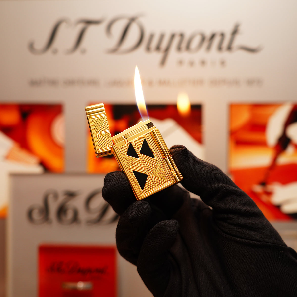 A gloved hand holds a lit Vintage 2000 St Dupont Ligne 2 Lighter in 24k Gold Finish with Africa Natural Lacquer against a blurred S.T. Dupont branded background.