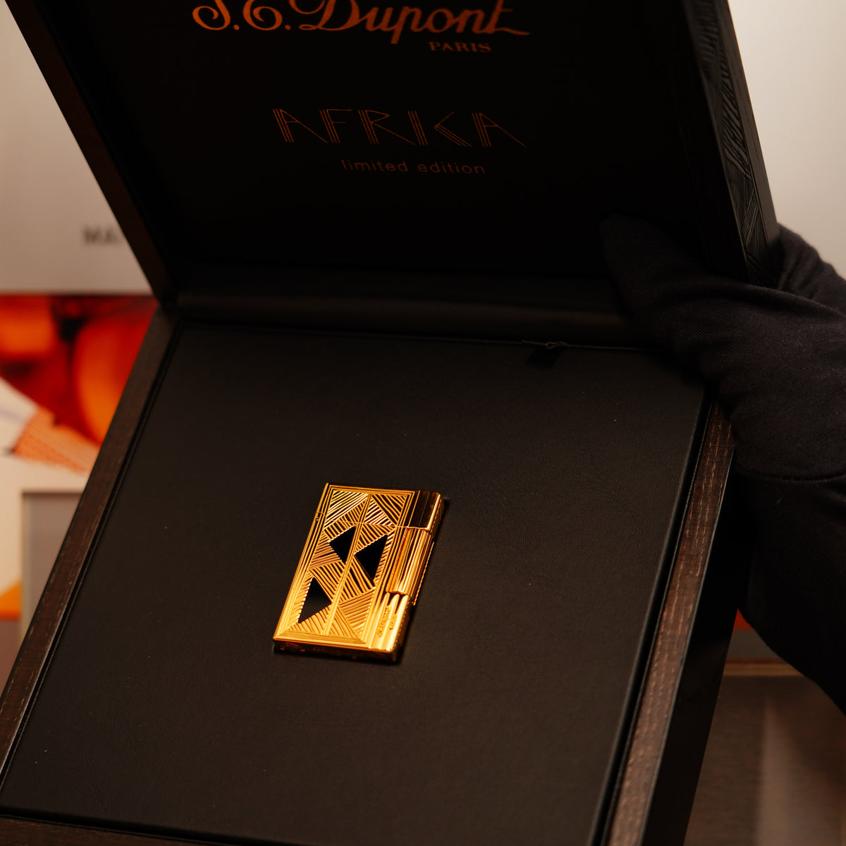 A 24k gold finish S.T. Dupont Afrique Limited Edition Ligne 2 lighter with geometric patterns is displayed in an open black box, held by a gloved hand.