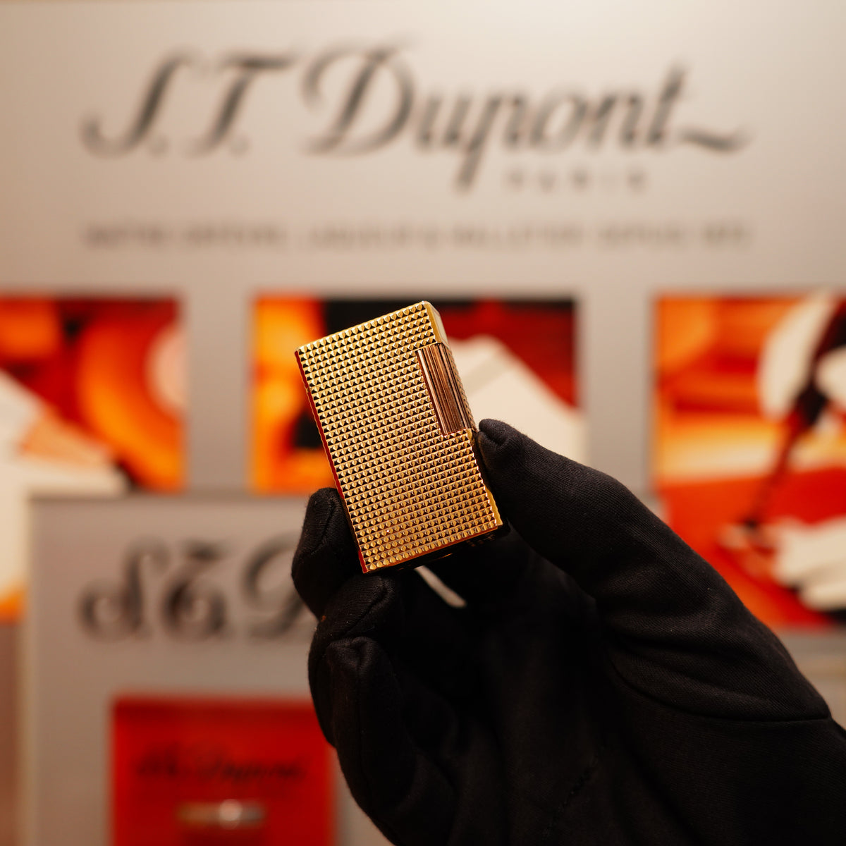 A black-gloved hand holds a Vintage 1990 S.T. Dupont 24k Gold Iconic Ligne 1 Diamond Head Pattern Lighter against a blurred background featuring "S.T. Dupont" and various colors.