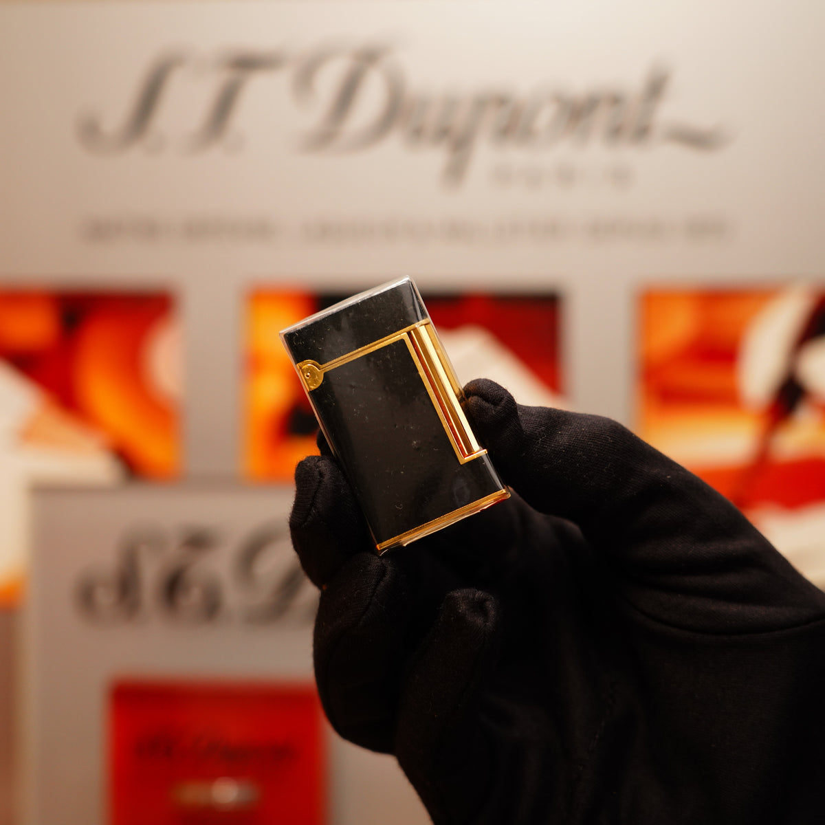 A gloved hand holds a Vintage 1990 S.T. Dupont Overall Black Lacquer Early 24k Gold Ligne D Sealed Lighter against a blurred background displaying the "S.T. Dupont" logo.