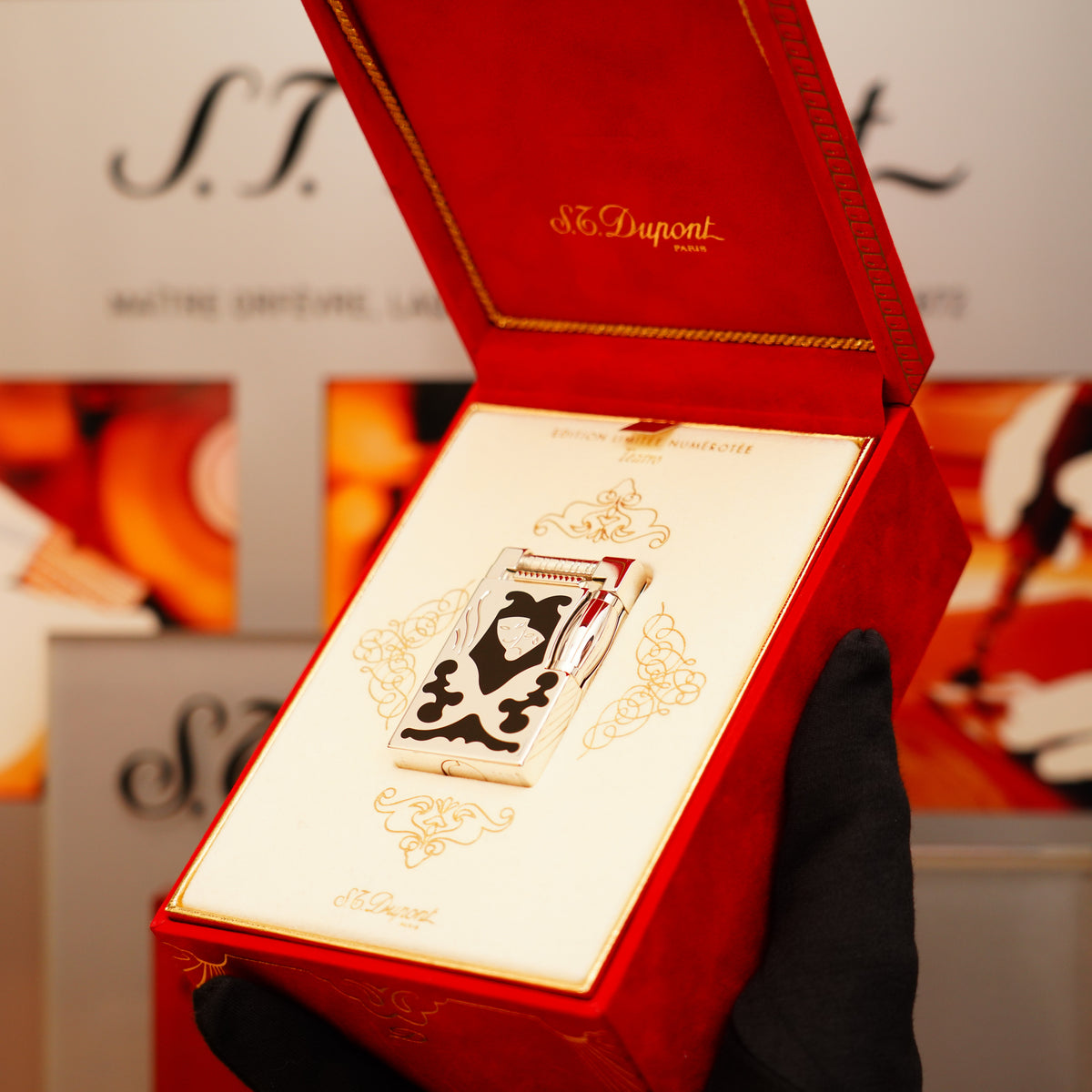 A gloved hand holds a Vintage 1997 St Dupont Heavy Platinum Black Natural Lacquer Early Limited Edition S.T. Dupont lighter, displayed in a red velvet presentation box with an ornate white and black design.