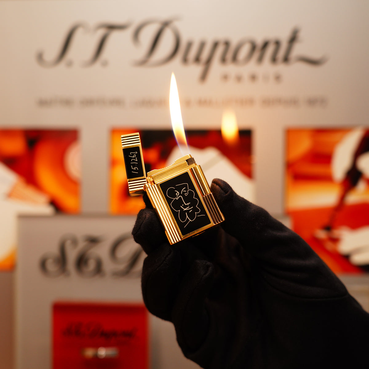 A gloved hand holds a lit S.T. Dupont Vintage 1990 Important St Dupont 24k Gold finish Pablo Picasso Limited Edition Ligne 1 black lacquer Lighter; blurred signage appears in the background.
