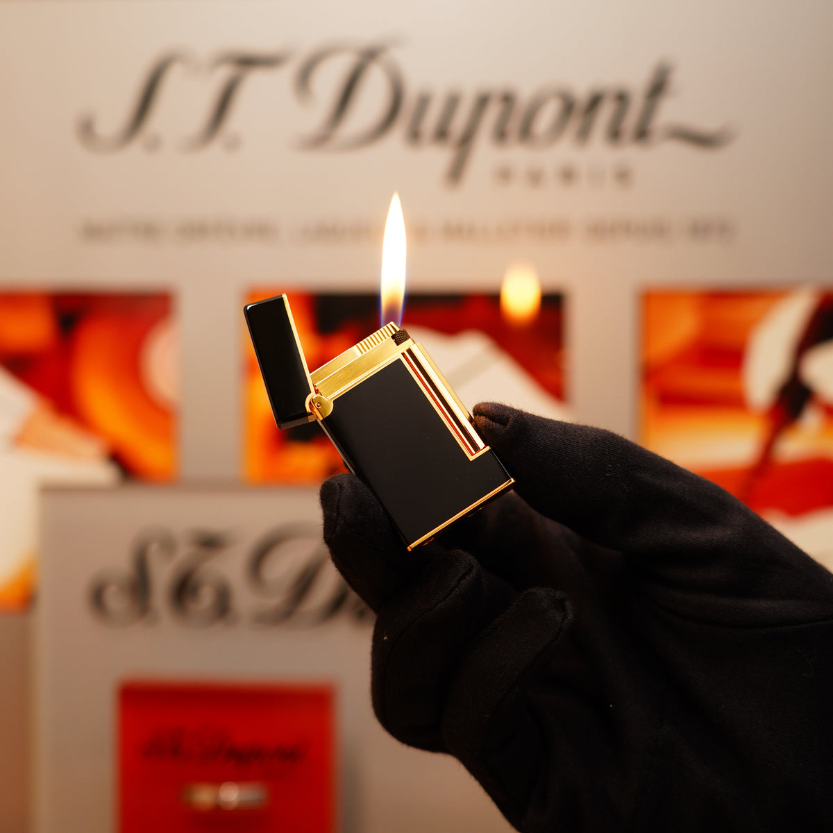 A gloved hand holds a lit Vintage 1990 St Dupont Overall Black Lacquer Early 24k Gold Ligne D Lighter against a blurred S.T. Dupont Paris background.