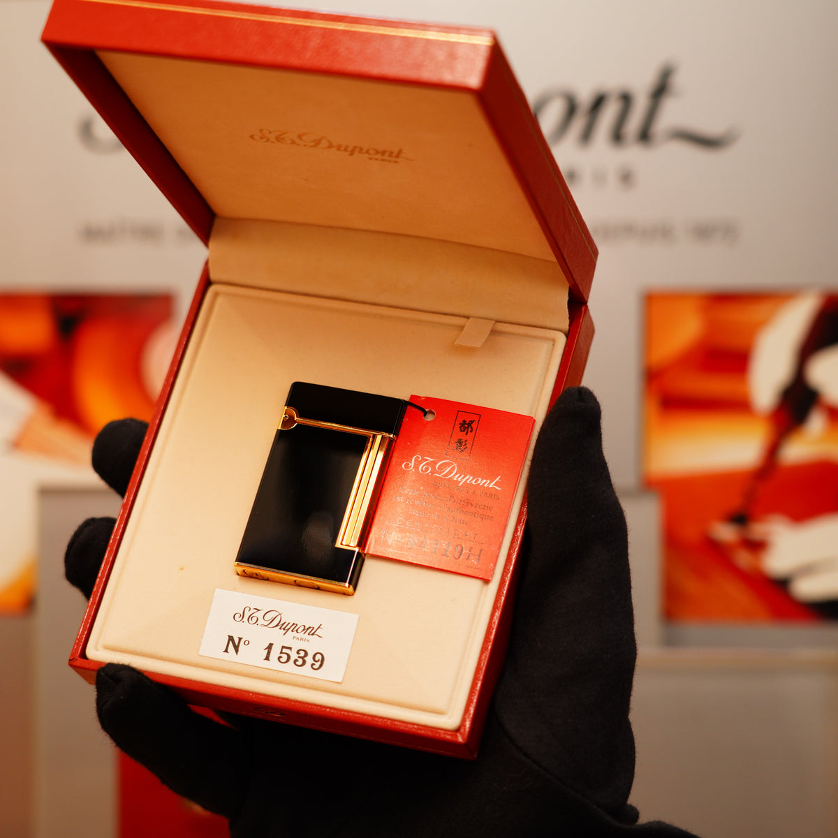 A gloved hand holds a Vintage 1990 S.T. Dupont Overall Black Lacquer Early 24k Gold Ligne D Lighter in an open red box with a label marked "No 1539.