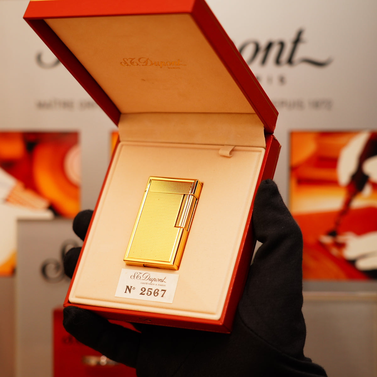 A black-gloved hand holds an open red box showcasing a gold Vintage 1990 S.T. Dupont 24k Large Grain d'Org Iconic Ligne 1 Heavy Finish Lighter, numbered 2567, against a blurred background.