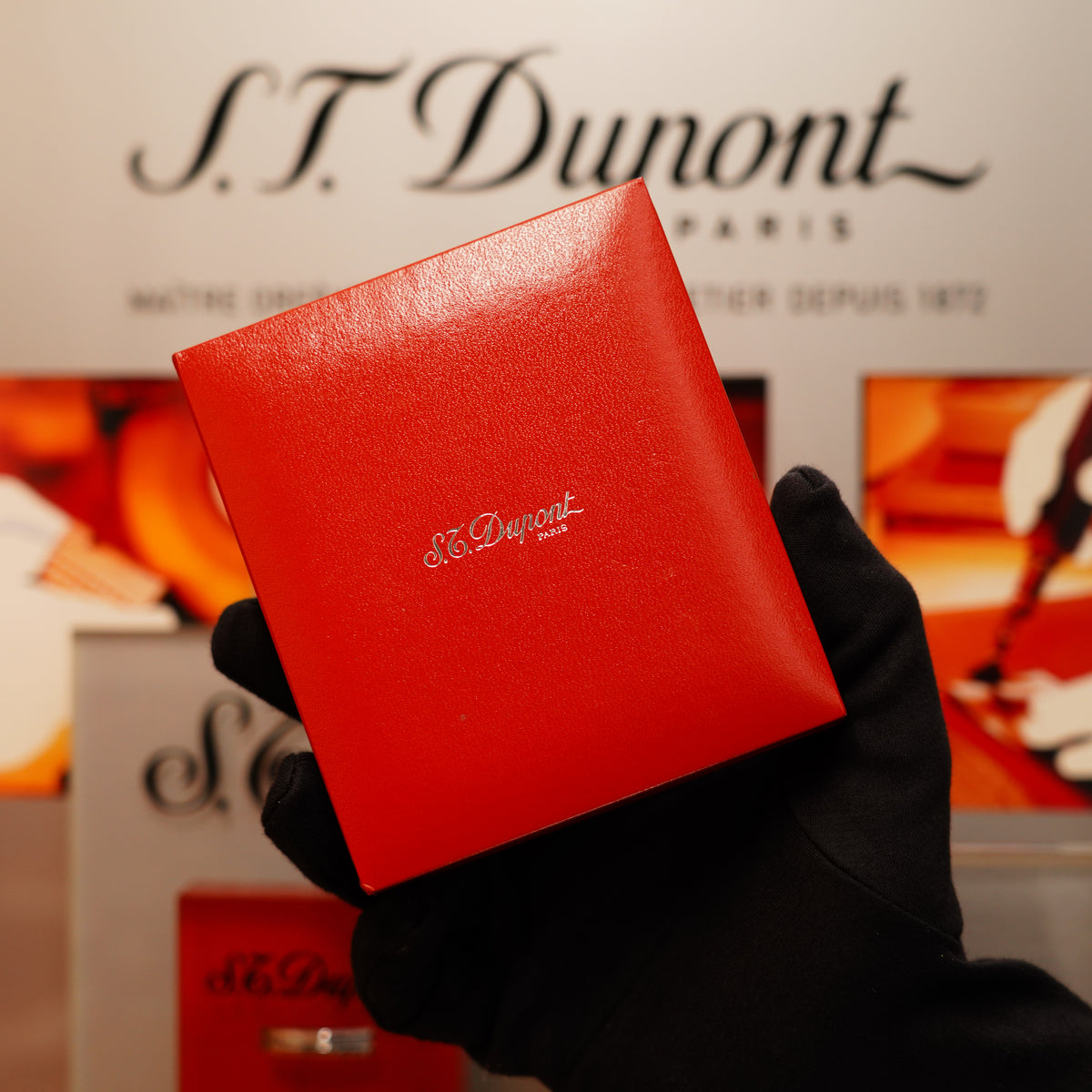 A person in black gloves holds a red S.T. Dupont Paris box containing a Vintage 2000 St Dupont Heavy Silver Finish Small iconic Ligne 1 Vertical Ligne Lighter, with the brand's logo and promotional materials in the background.