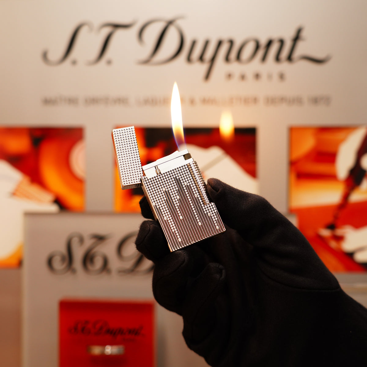 A gloved hand holds the lit Vintage 2005 S.T. Dupont Rare Natural Diamond Drops Large Ligne 2 Heavy Platinum Limited Edition Lighter against a backdrop featuring the S.T. Dupont logo and branded display graphics.