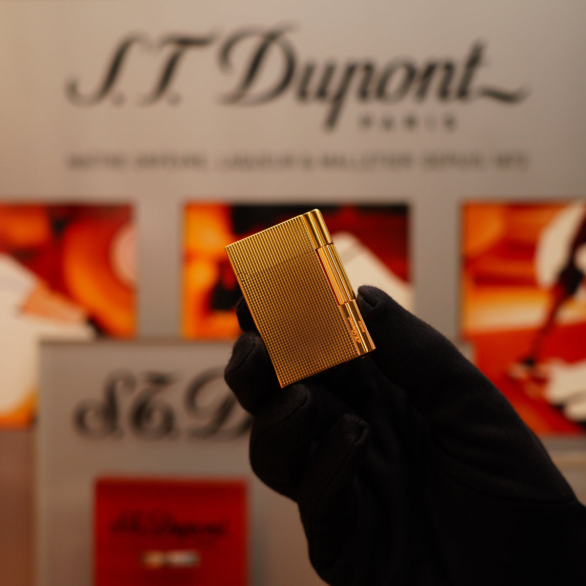 A gloved hand holds a Vintage 2002 S.T. Dupont Rare Heavy 24k Gold Finish Gatsby Ligne 2 Lighter with a vertical line pattern against a backdrop featuring the brand's logo and blurred red and white elements.