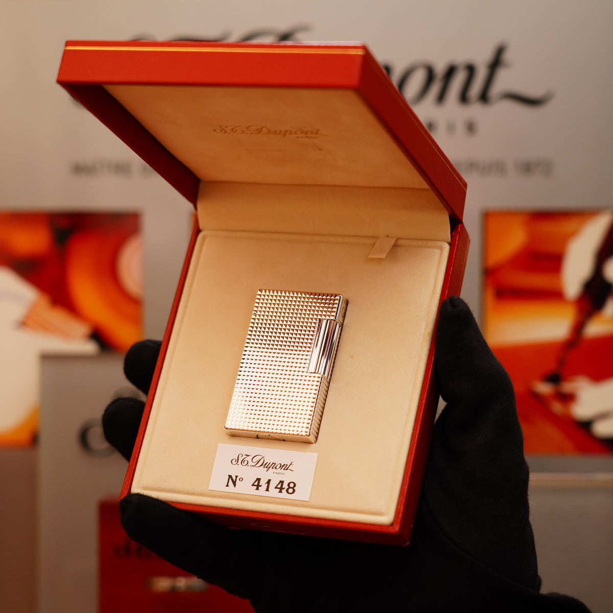 A gloved hand holds an open red box displaying a "Vintage 1996 St Dupont Ligne 1 Diamond Head Pattern Large Heavy Silver Finish Lighter" by S.T. Dupont inside a store.
