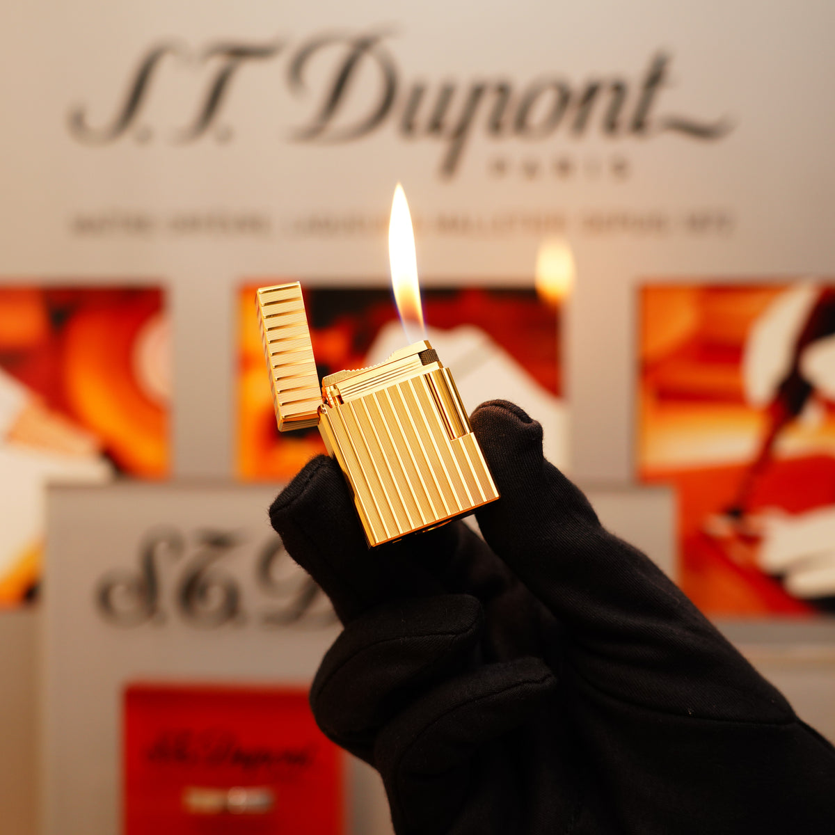 A gloved hand holds a Vintage 1990 S.T. Dupont 24k Gold Finish small Ligne 1 rare Pattern Lighter with a flame, against a blurred S.T. Dupont Paris display background.