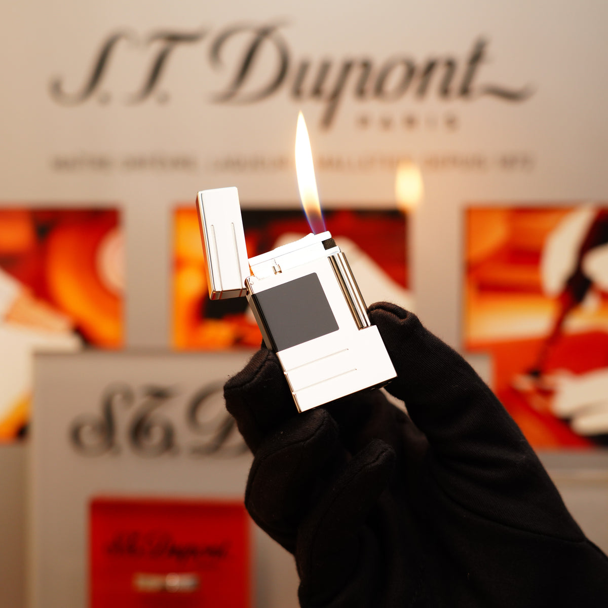 A gloved hand holds an open flame from a Vintage 1999 St Dupont Abstraction Ligne 2 Black Lacquer Lighter against a blurred S.T. Dupont background.