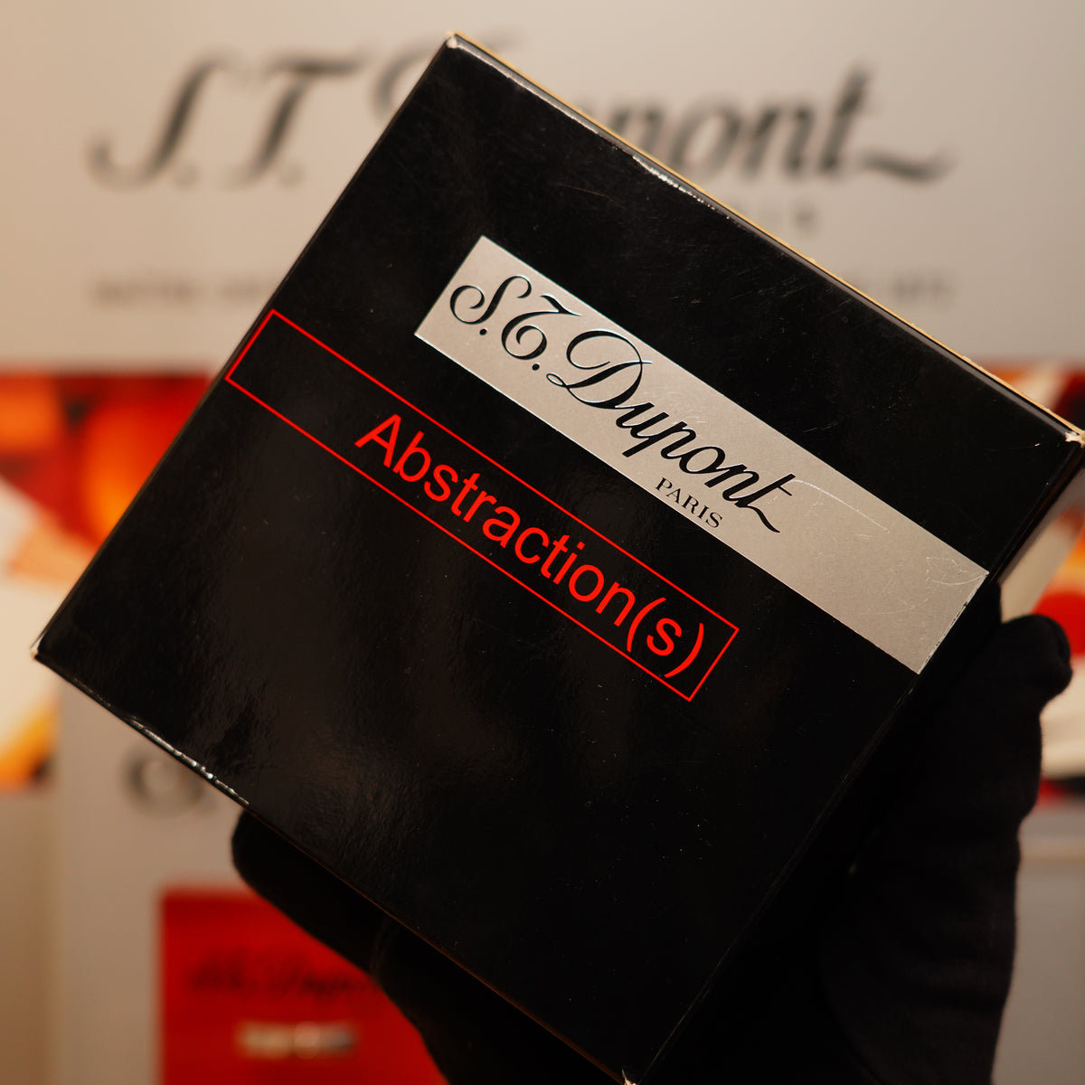 A gloved hand holds a black box labeled "S.T. Dupont," featuring the Vintage 1999 St Dupont Abstraction Limited to 2500 Heavy Palladium Finish Ligne 2 Black Lacquer Lighter, against a blurred background.