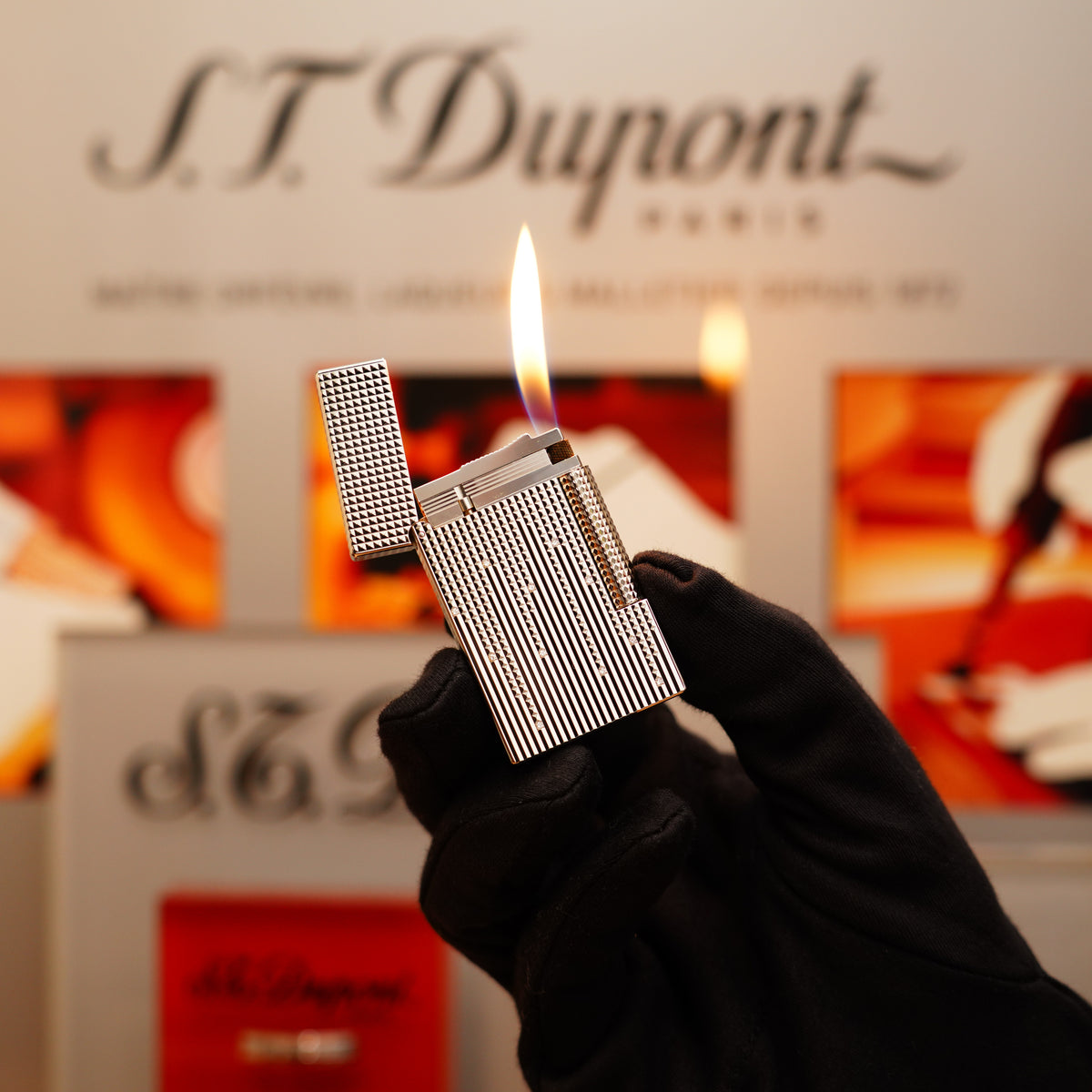 A gloved hand holds a lit metal lighter, part of the S.T. Dupont Vintage 2005 Diamond Drops Heavy Platinum Limited Edition Ligne 2 collection, featuring 22 natural diamonds, against a blurred background showcasing the brand's logo.