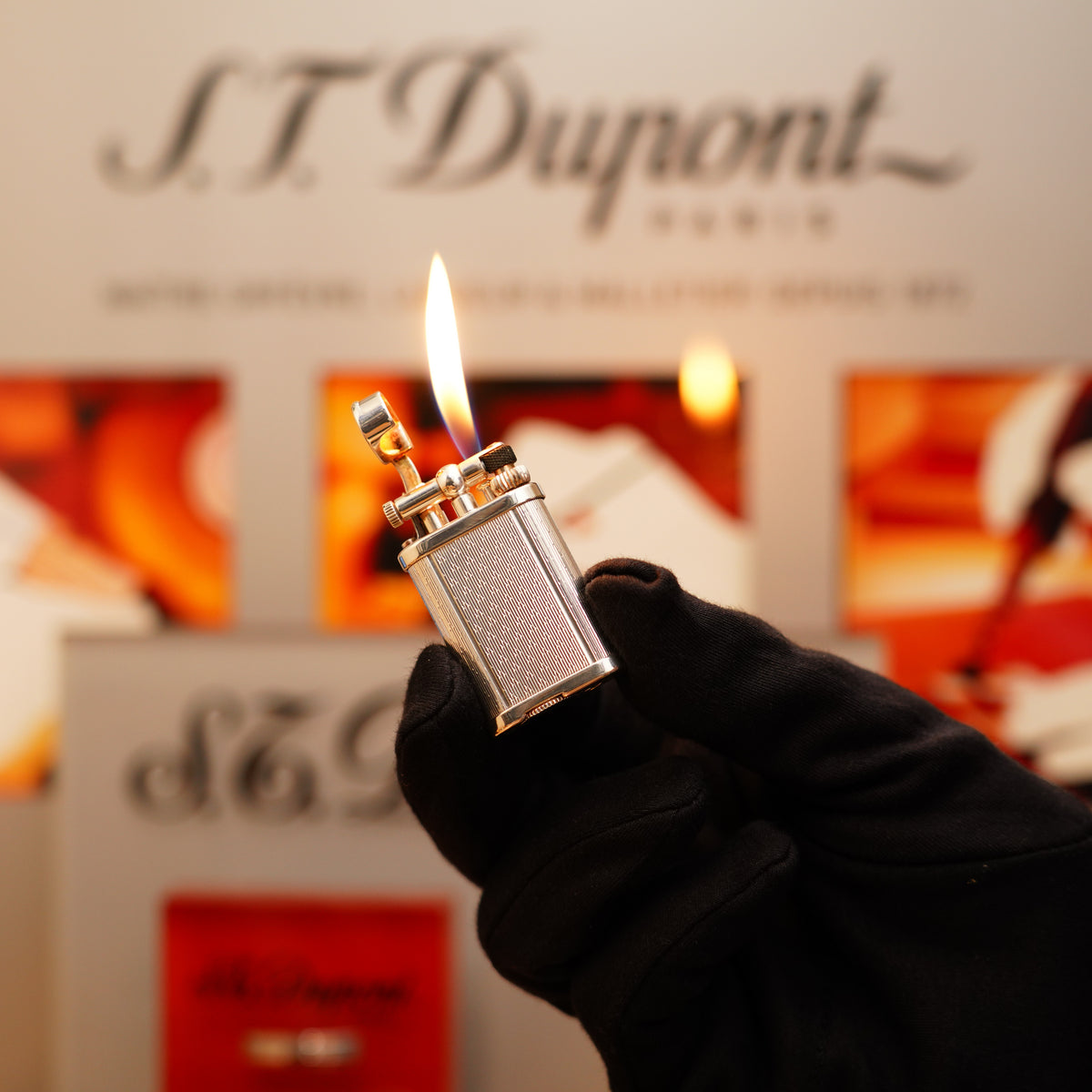 A gloved hand holds a lit Vintage 1990 Dunhill mini Unique Heavy Silver Finish Rare pattern Lighter with a flame, in front of a blurred S.T. Dupont logo in the background.