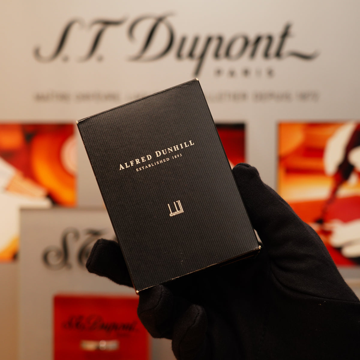 A person wearing a black glove holds a box of the Vintage 1990 Dunhill Mini Unique Heavy Silver Finish Rare Pattern Lighter, with a blurred S.T. Dupont Paris backdrop.