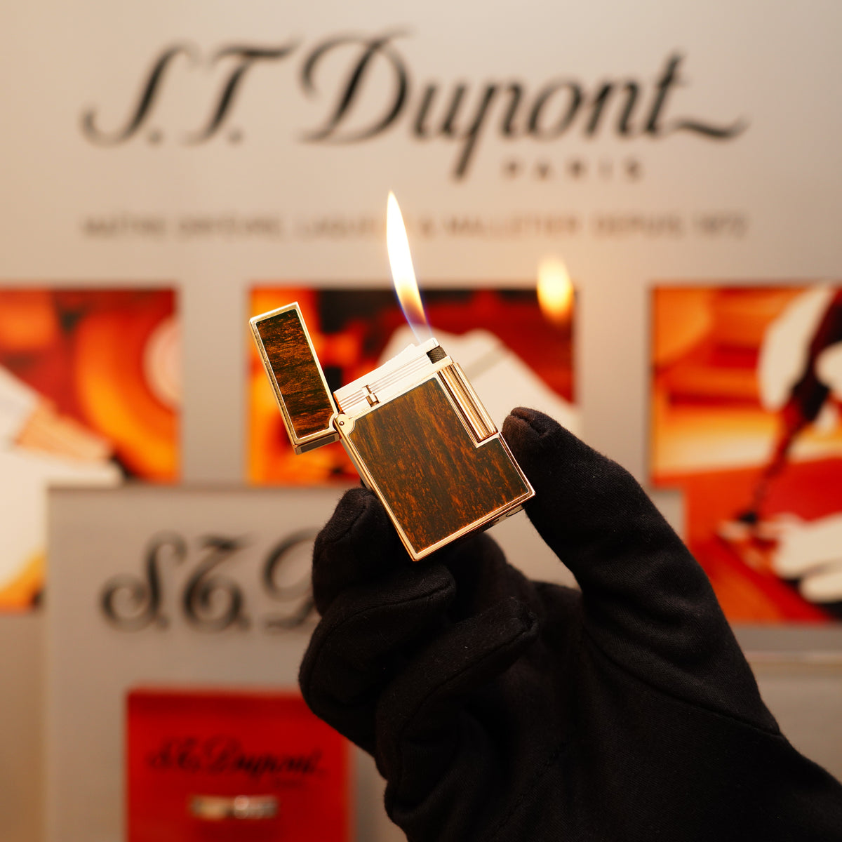 A gloved hand holds an open Vintage 1970 S.T. Dupont Ligne 2 lighter in heavy gold and wood finish, with a lit flame against a blurred background displaying the brand name.