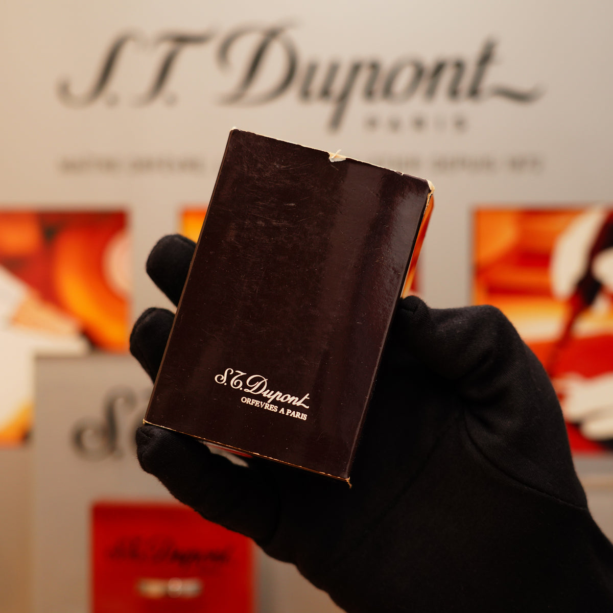 A hand in a glove holds a brown box branded with "S.T. Dupont," featuring the Vintage 1970 St Dupont 24k Heavy Gold Finish Real Wood Medium Ligne 2 Natural Lighter, against a blurred backdrop with more S.T. Dupont logos.