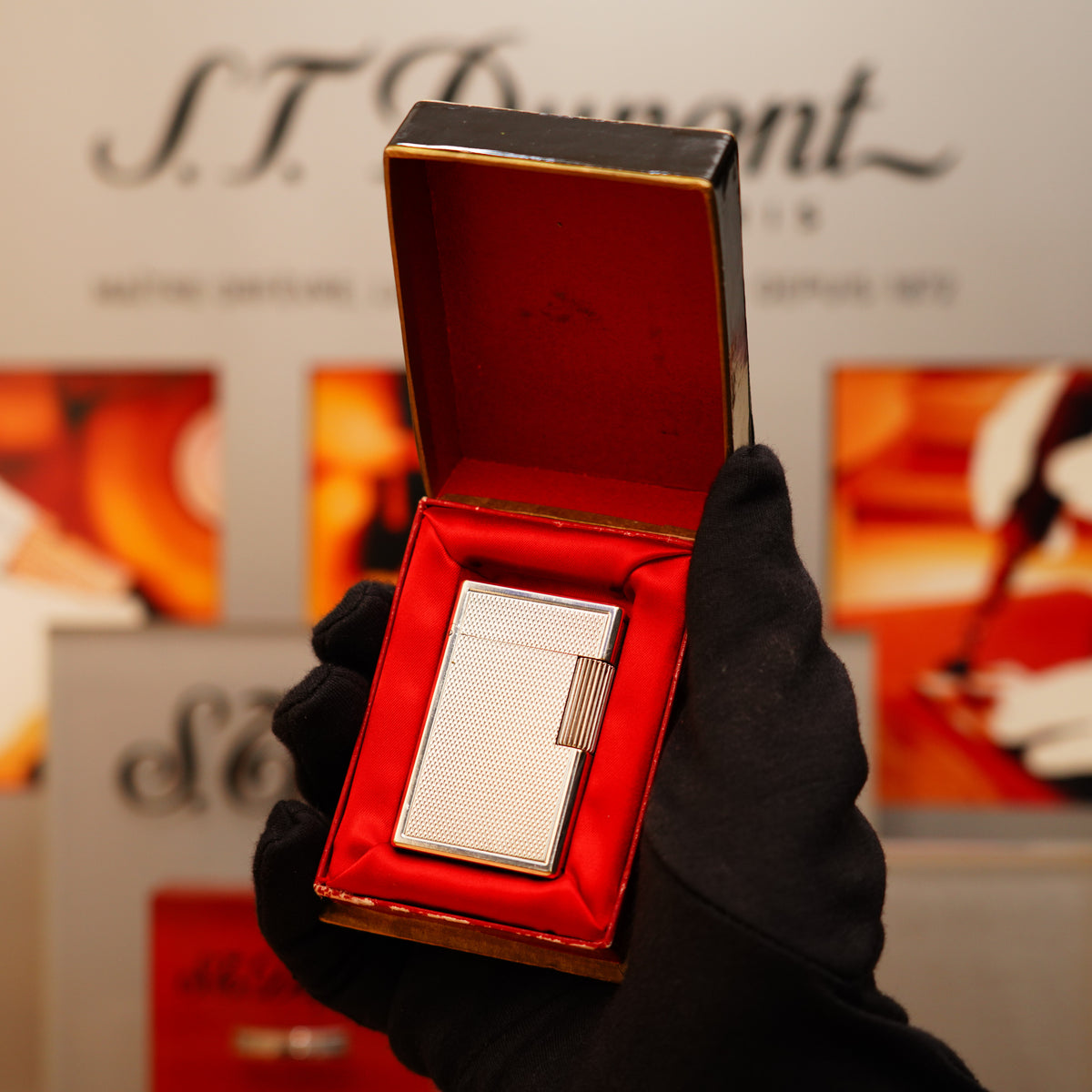 A person wearing black gloves holds an open box with a Vintage 1940 St Dupont Petrol initial version Essence Heavy Silver Finish Extremely rare condition lighter inside, against a blurred background with S.T. Dupont signage.