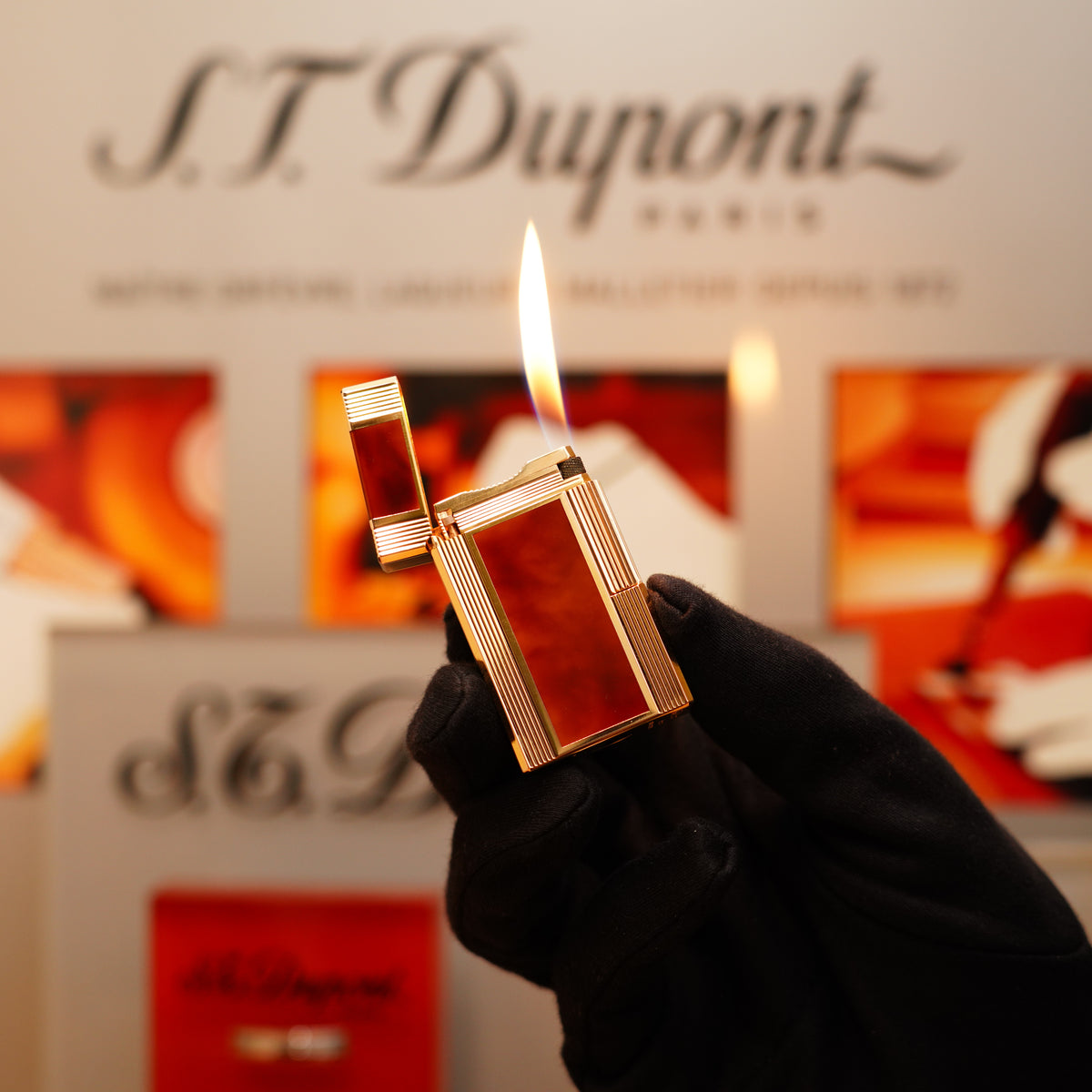 A hand in a glove holds a lit Vintage 1970 S.T. Dupont Japanese Lacquer Signed 24k Gold Large Ligne 1 Lighter with a natural mid-brown lacquer design.