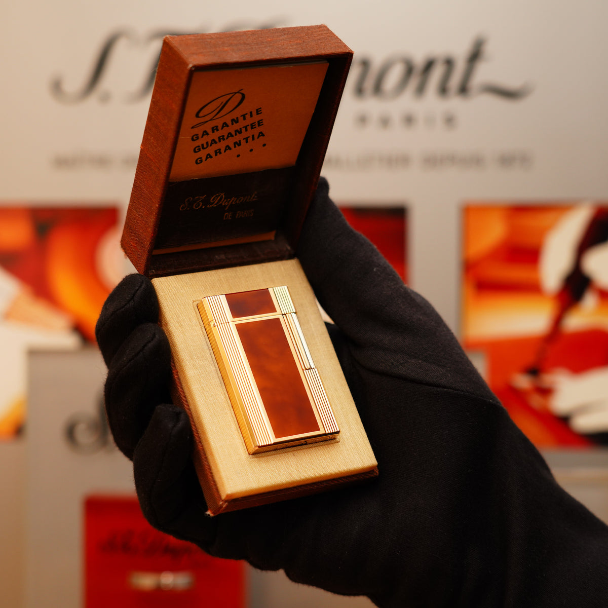 A gloved hand holds a Vintage 1970 S.T. Dupont Ligne 1 model lighter with mid-brown lacquer and heavy gold, displayed in an open box against a blurred backdrop showing the brand name.
