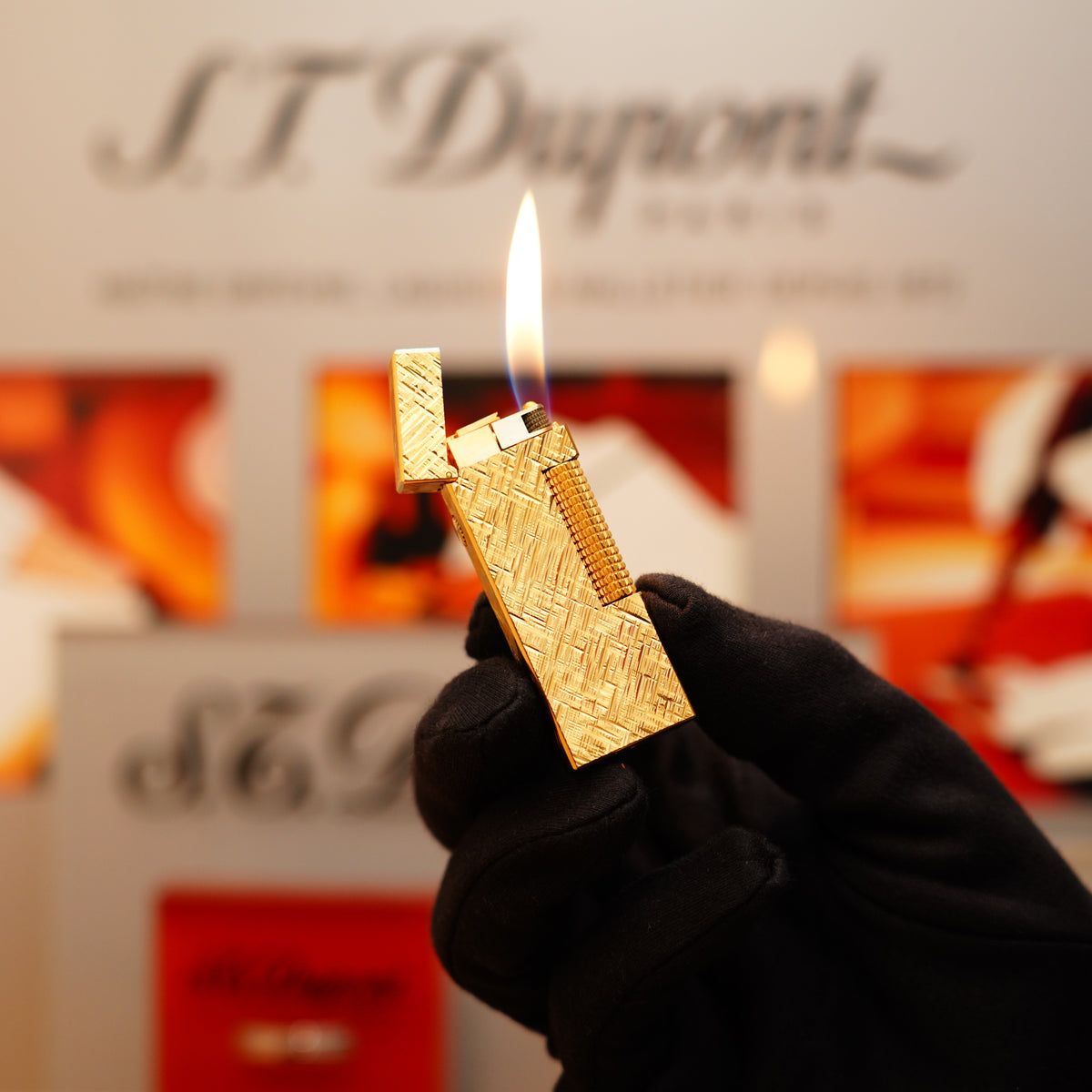 A gloved hand holds a lit Vintage 1975 Dunhill 24k Gold Finish Rollagas Lighter, showcasing its intricate design against a blurred background featuring S.T. Dupont branding.