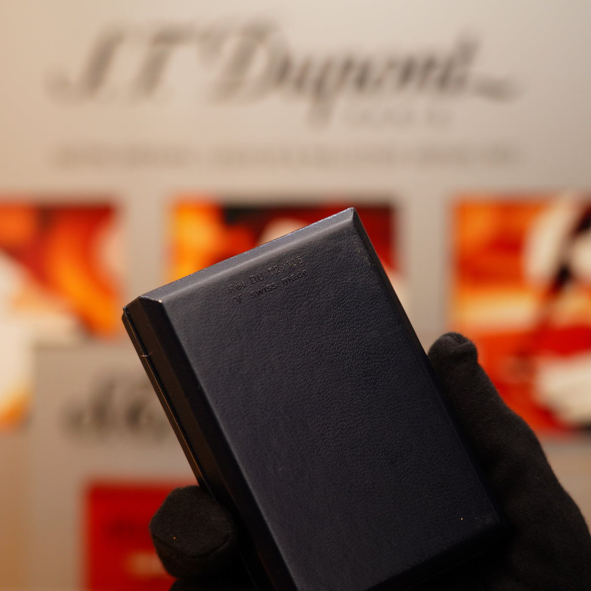 A gloved hand holds a black Dunhill item against a blurred background of S.T. Dupont branding.