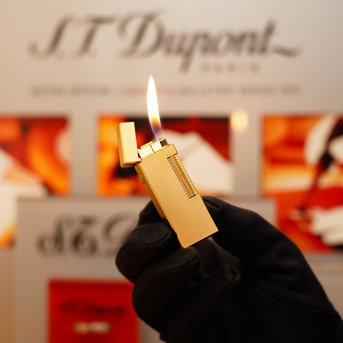 A gloved hand holds a lit Vintage 1970 Dunhill 24k Gold Finish Rollagas Diamond Head Pattern Initial Version Lighter, with a blurred branded background.