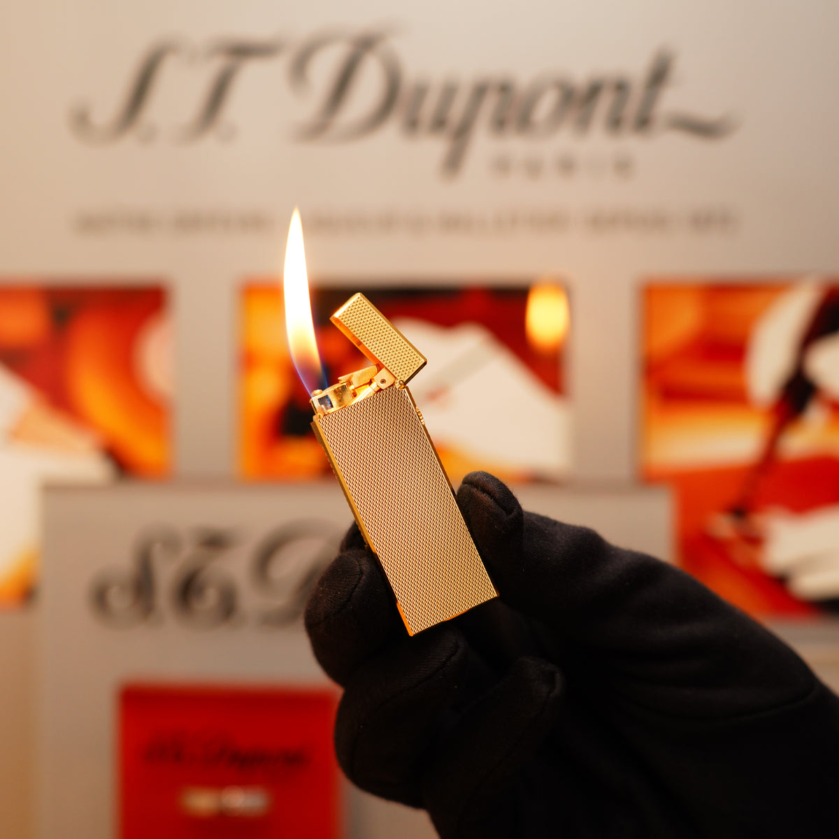 A gloved hand holds a Vintage 1971 Dunhill 24k Gold Finish Rollagas Barley Pattern Initial Version Lighter with a visible flame against a blurred backdrop featuring the S.T. Dupont logo.