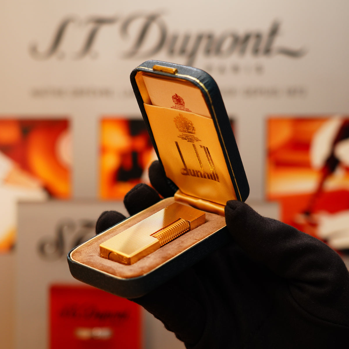 A gloved hand holds an open velvet-lined box containing a Vintage 1971 Dunhill 24k Gold Finish Rollagas Barley Pattern Initial Version Lighter. The background features indistinct signage and blurred images.