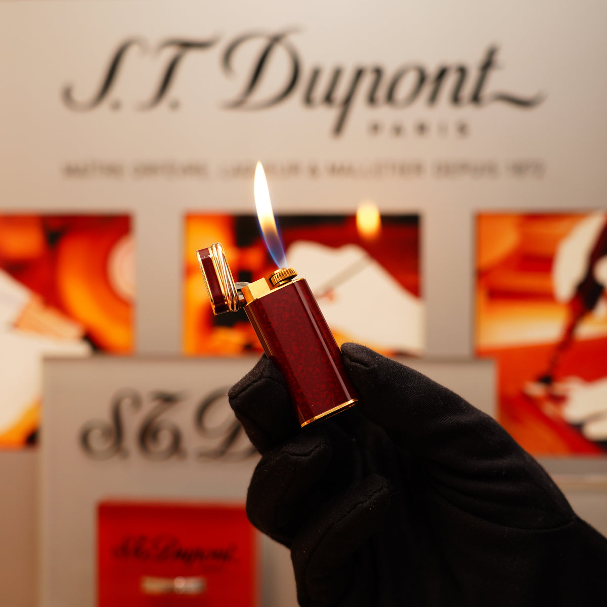 A gloved hand holds a lit Vintage 1980 Cartier Mini Pentagon Red Marble Lighter with 24k Gold Finish and 18k Trinity Ring in front of branded packaging.