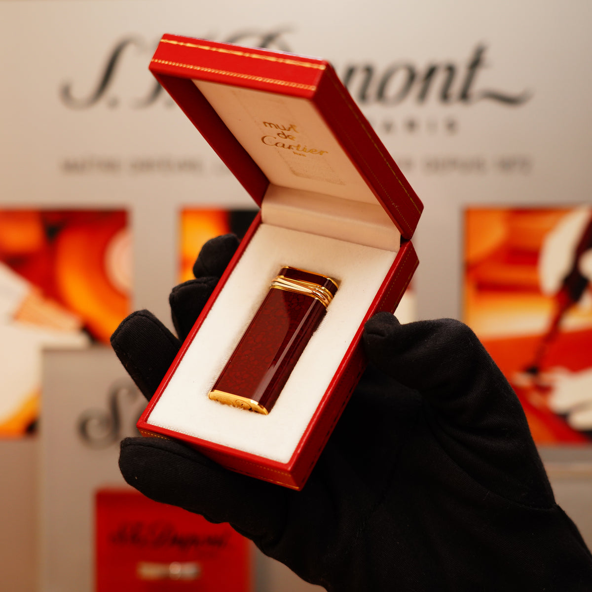 A gloved hand holds an open red box displaying a shiny Vintage 1980 Cartier Mini Pentagon lighter in red marble and 24k gold finish, with "must de Cartier" inscribed inside the lid.