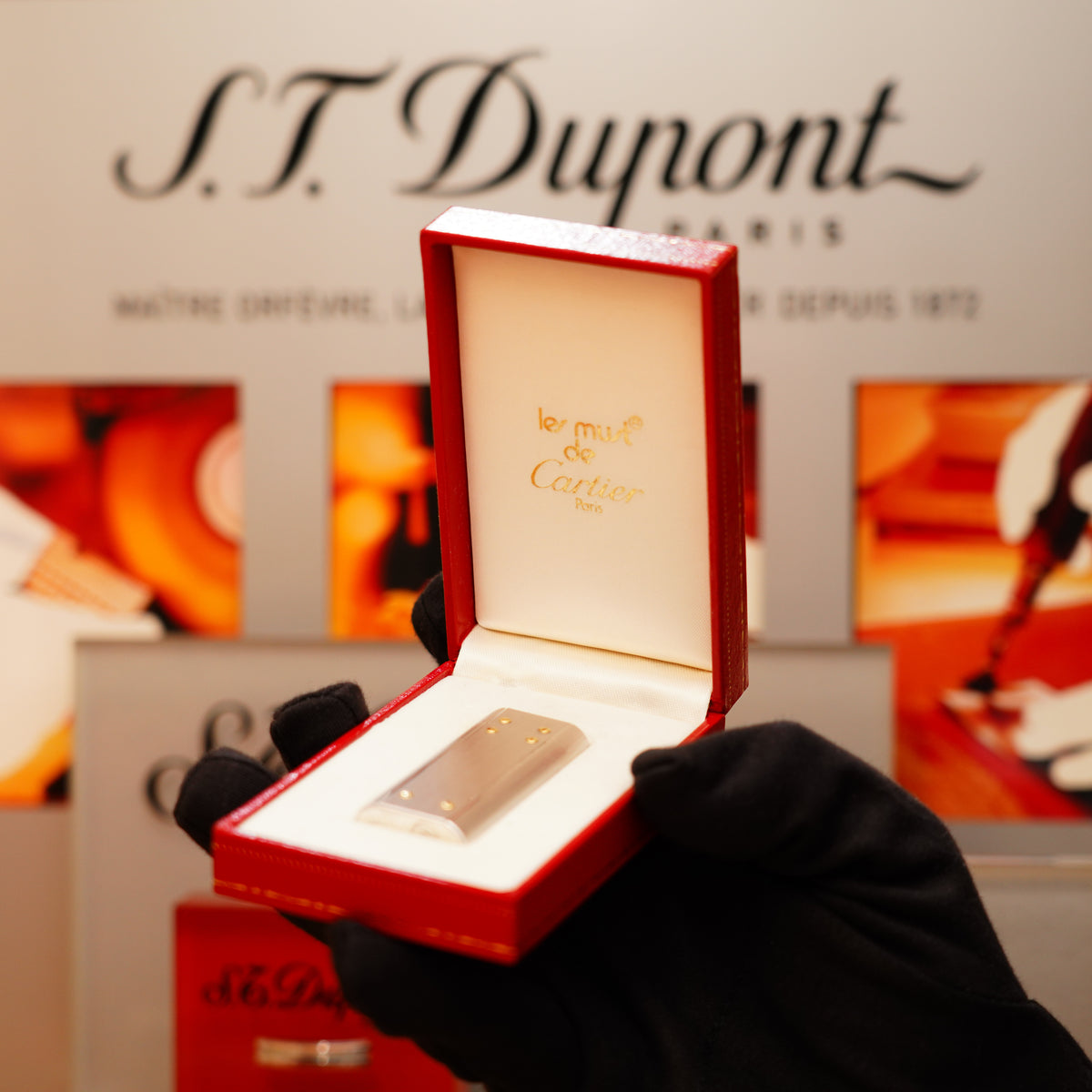 A hand in a black glove holds an open red Cartier box revealing a Vintage 1980 Cartier Mini Iconic Santos Paladium Finish with 18k screw lighter, in front of an S.T. Dupont sign.