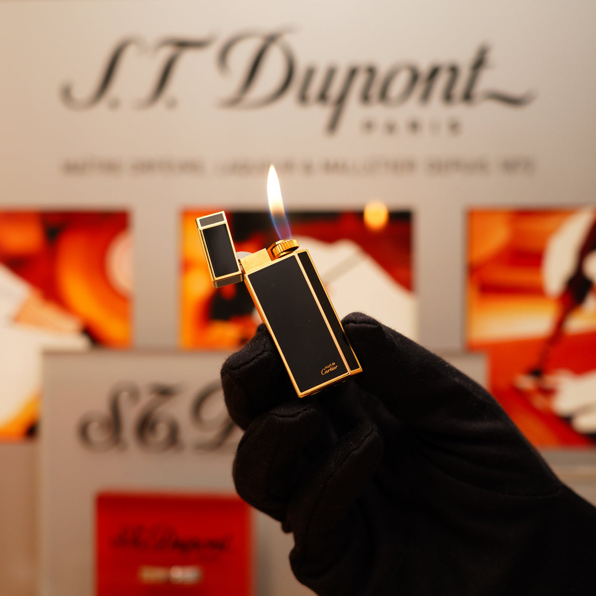 A gloved hand holds a lit Vintage 1980 Cartier Mini Pentagon Lighter, featuring a 24k gold finish and full black natural lacquer, in front of branded signage.
