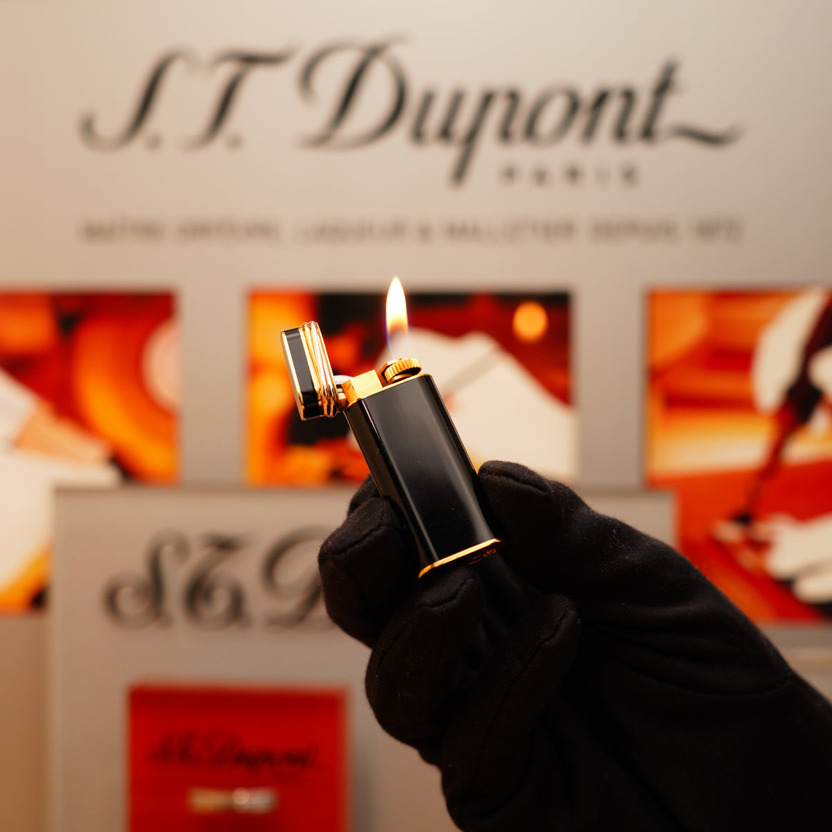 A gloved hand holds a lit rare Vintage 1980 Cartier Mini Pentagon lighter with a full black 24k gold finish against a blurred branded backdrop.