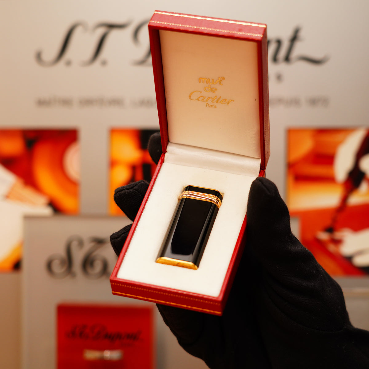 A hand in a black glove holds an open red box with a rare Cartier vintage 1980 mini pentagon lighter, featuring a full black 24k gold finish and 18k Trinity ring. The lid of the box reads "Cartier Paris.