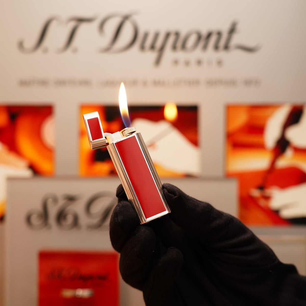 A gloved hand holds a lit Vintage 1970 Cartier solid sterling red enamel lighter against a blurred background showing the Cartier logo.