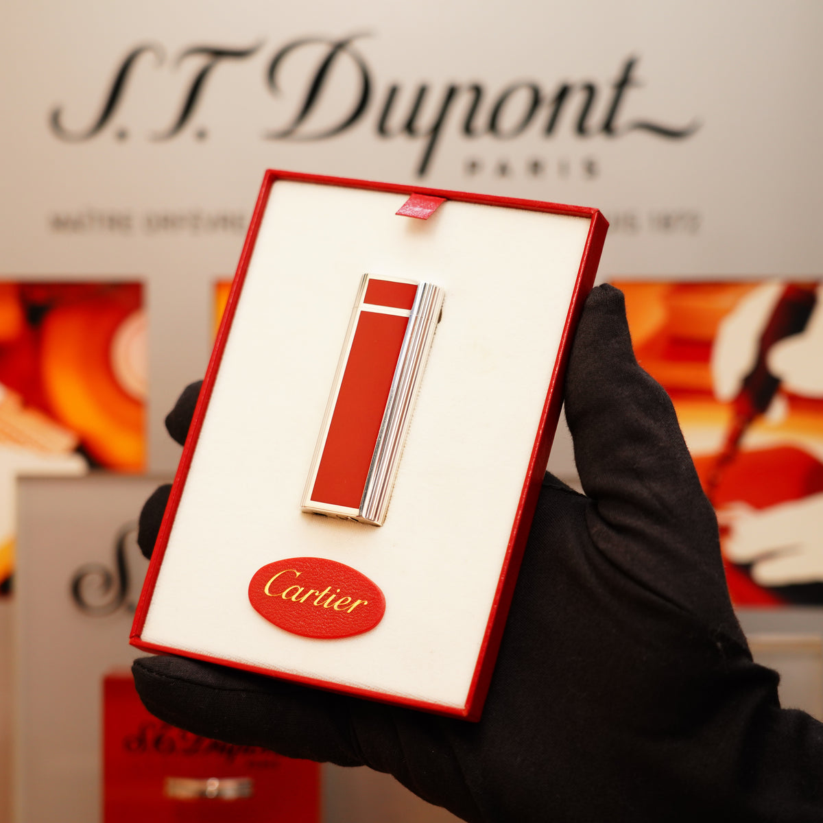 A gloved hand holds a Vintage 1970 Extremely Rare Cartier Solid Sterling Red Enamel Solid Silver 925 Hallmark lighter displayed in a box, with blurred Cartier branding in the background.