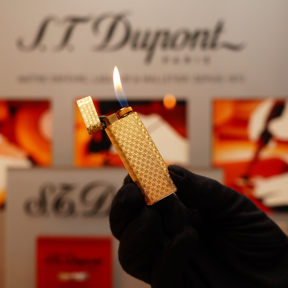 A hand in a glove holds the Vintage 1970 Extremely Rare Cartier Solid 18k Gold Visual Guilloché Pattern Lighter aflame, set against a background of blurred Cartier branding and abstract art.