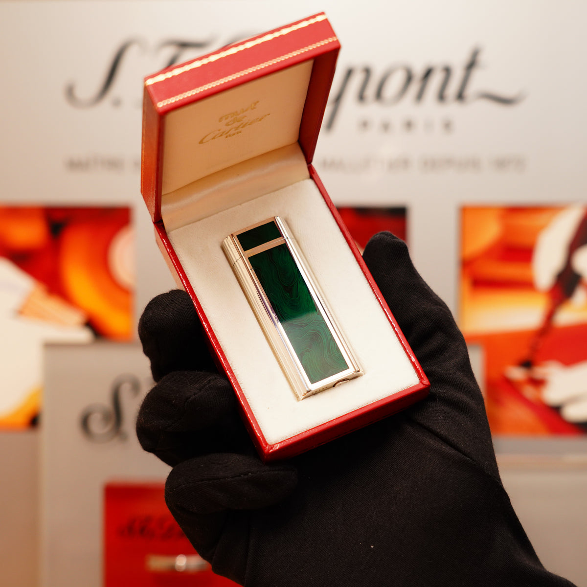 A hand in a black glove holds the Vintage 1970 Extremely Rarest Cartier Solid Sterling Green Enamel Solid Silver 925 Hallmark lighter in an open red box with a white interior, against a background showing a blurred Cartier display.