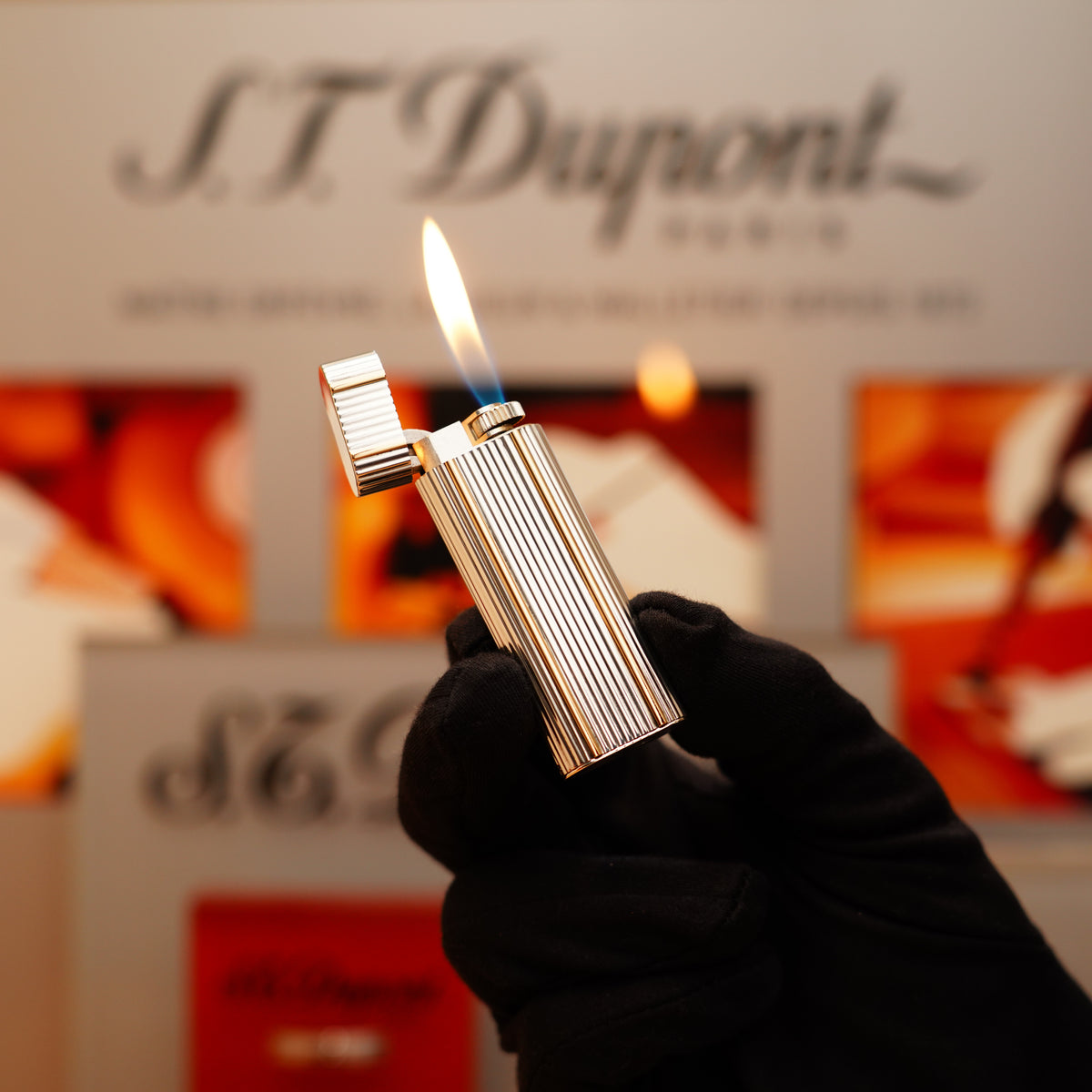 A gloved hand holds a lit Vintage 1970 Cartier Sterling lighter with 18k lines against a blurred background of "S.T. Dupont" signage.