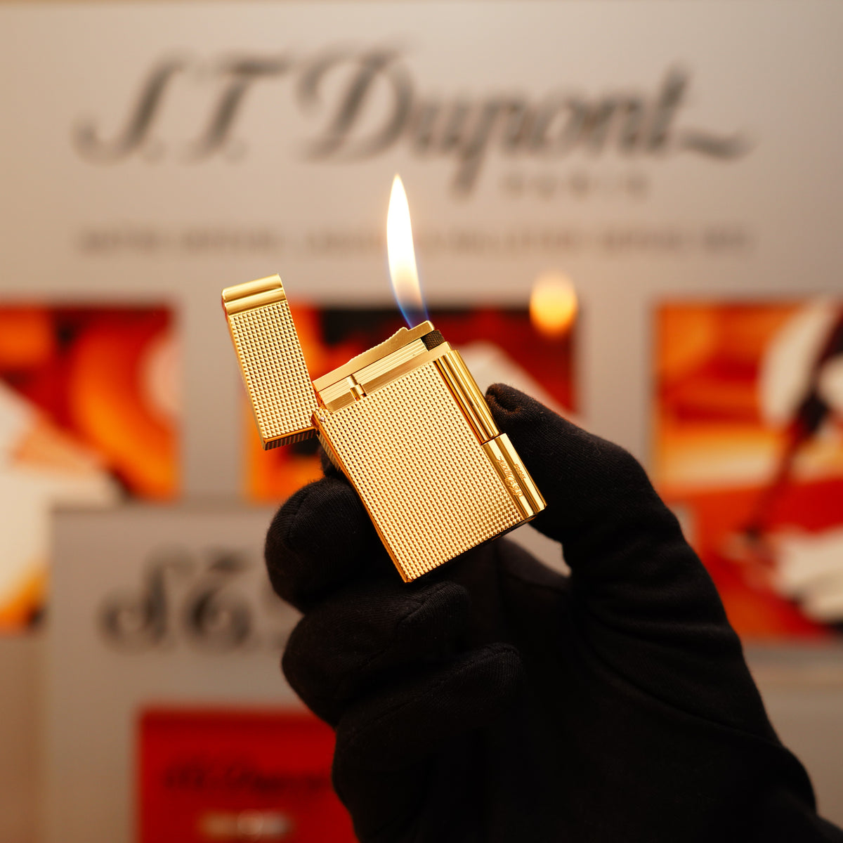 A gloved hand holds a lit, gold-colored Cartier lighter featuring a textured diamond pattern, against a blurred background with "S.T. Dupont" text.