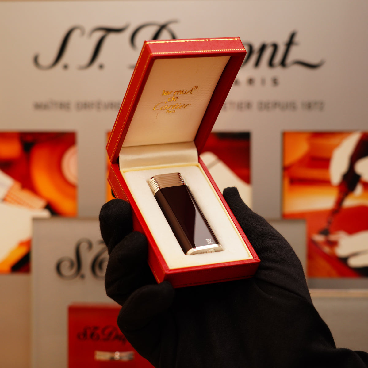 A gloved hand holds an open red box displaying a sleek, Vintage 1970 Cartier solid 925 sterling silver lighter. Blurred branding and promotional images serve as the background.
