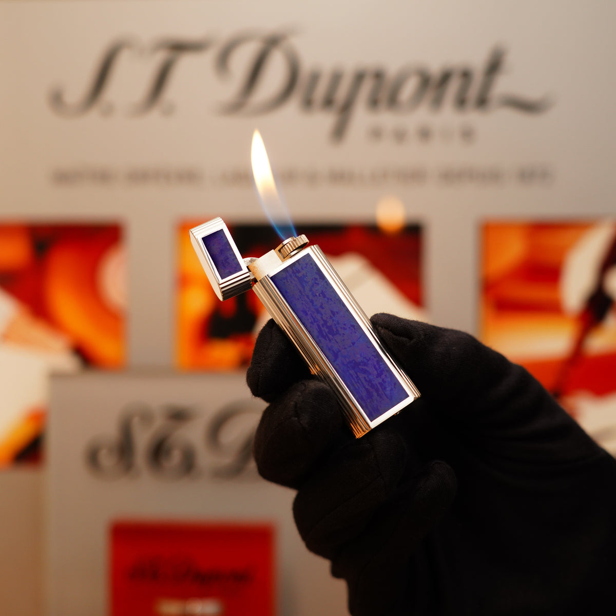 A person wearing a black glove holds a lit, metallic Cartier lighter featuring blue guilloché enamel. The S.T. Dupont logo is visible in the blurred background.