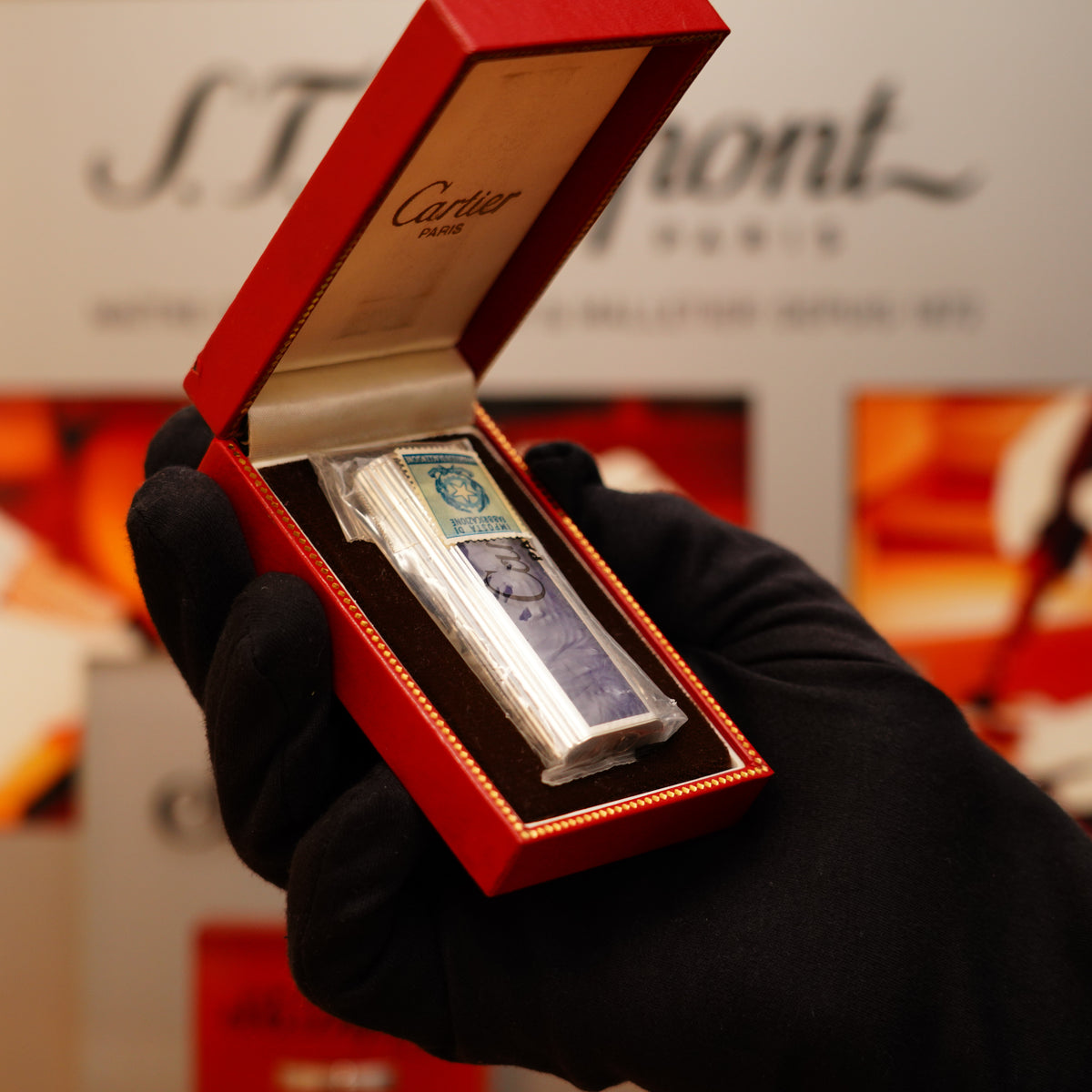 A hand in a black glove holds an open red box featuring the Vintage 1970 Extremely Rare Cartier Sterling Blue Guilloché Enamel Solid Silver 925 Hallmark factory lighter, an ornate silver item with intricate designs and a blue detail on top.