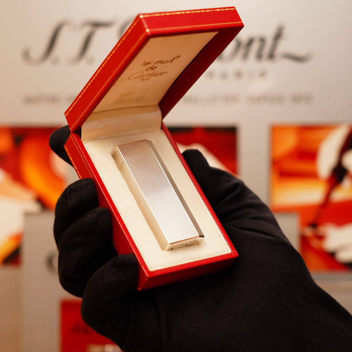A hand in a black glove holds an open red box containing a Vintage 1980 Cartier Heavy Silver Finish Pentagon Lighter.
